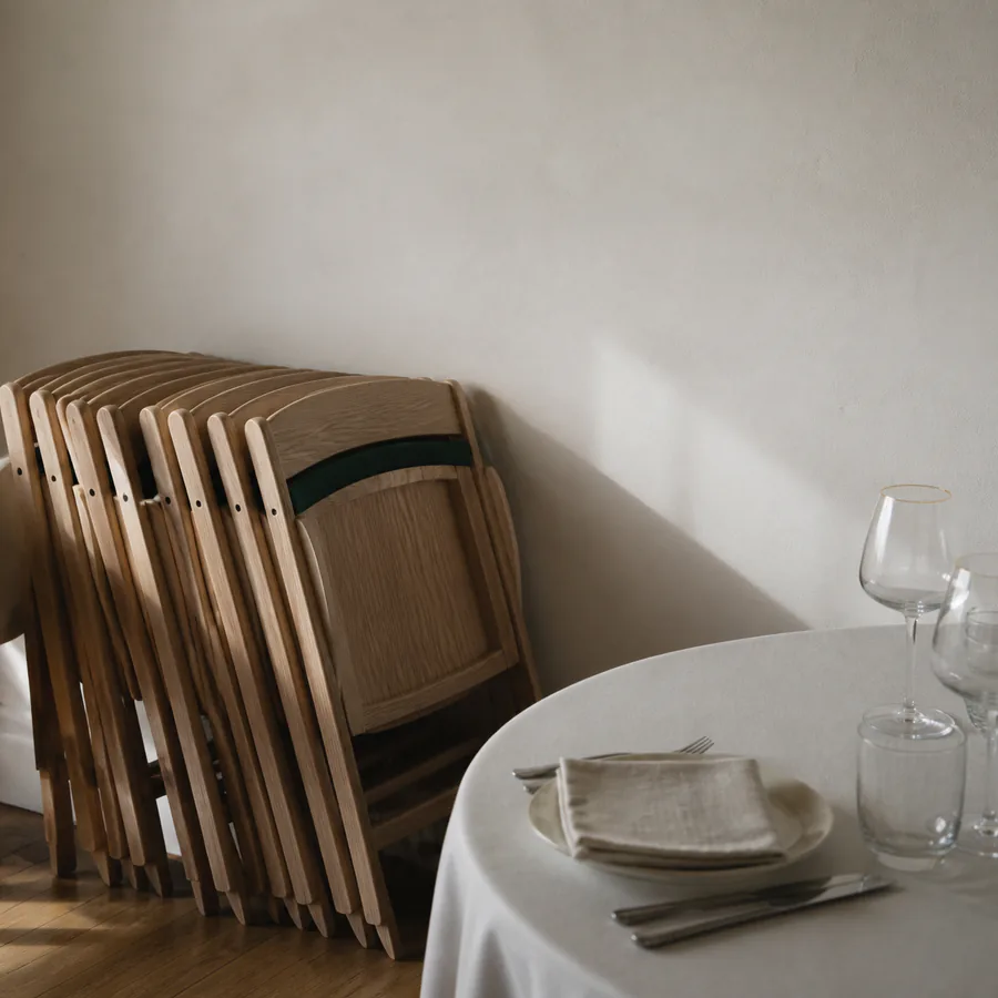 A stack of pale wooden folding chairs against a plain plaster wall in an empty venue, beside a round table laid with a white cloth, linen napkin and glassware.